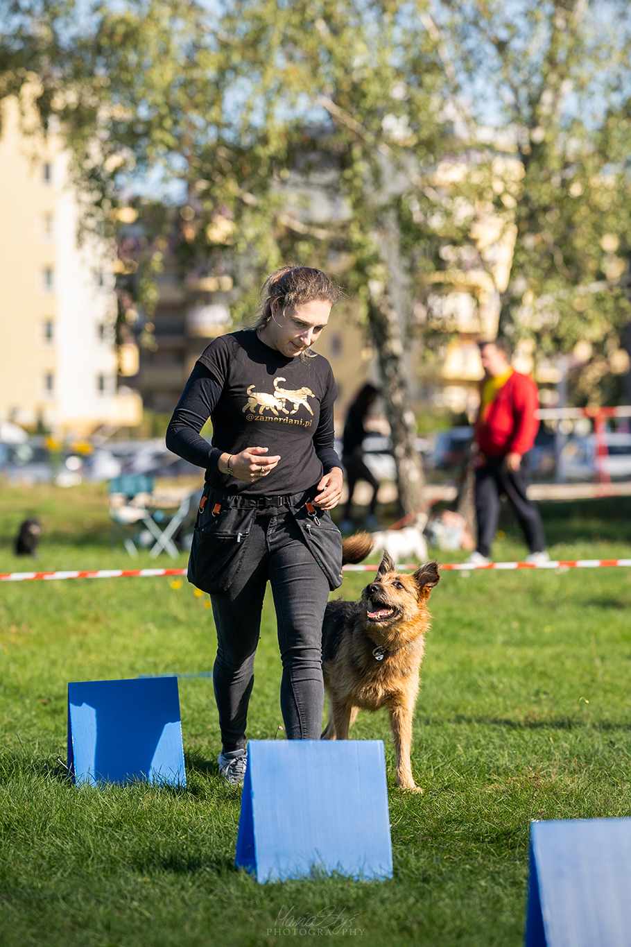 Rally-Obedience — czyli, czy posłuszeństwo sportowe może być na luzie? Zdjęcie przedstawia kobietę trenującą psa na świeżym powietrzu, prawdopodobnie w ramach wydarzenia związanego ze szkoleniem psów. Kobieta ma na sobie koszulkę z logo "zamerdani.pl", a pies idzie obok niej, uważnie obserwując jej ruchy.